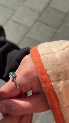A close-up shot shows a person's hand wearing a silver ring with a green, sparkly stone. The hand is holding another hand, which is slightly out of focus. The person wearing the ring has fair skin and a garment with an orange trim. The other person is wearing a black garment. The background shows a tiled pavement and sunlight. The hand with the ring rotates slightly, displaying the stone's glitter.
