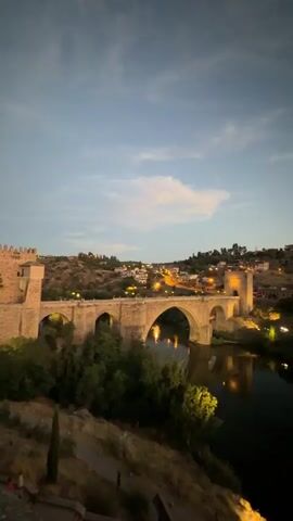 The video shows a wide shot of a stone bridge over a river at dusk. The bridge has multiple arches and is illuminated by artificial lights, casting reflections on the water. On the hill behind the bridge, there are buildings with lights on, suggesting a town or city. The sky is a dusky blue with some clouds. The surrounding landscape is hilly and covered with vegetation, with some trees visible along the riverbank. The camera appears to be stationary.