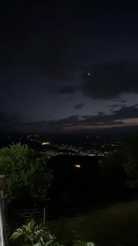 A camera pans across a dark outdoor scene at dusk or night. The foreground features a wooden bench and a white railing. Beyond the railing, a dark landscape with trees and a distant city illuminated by lights is visible. The sky is dark with visible clouds and some stars. In the middle ground, a dark silhouette of a statue of a person on a bicycle is visible behind the railing.
