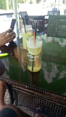 The video shows a close-up view of two glasses of lemonade on a dark, reflective table. The glass in the foreground is a jar with a logo that reads "CIMA Fruit Emotion". It contains lemonade with ice, lemon slices, and orange-colored straws. In the background, another glass of lemonade is visible. A person's hand holding a smartphone is in the upper left corner of the frame. To the left of the foreground glass, a yellow highlighter pen lies on the table. The environment appears to be an outdoor seating area, possibly a cafe or restaurant, with tables and chairs visible in the background and trees and foliage suggesting a natural setting. The lighting is bright, indicating daytime.