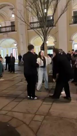 The video shows a group of people gathered in a public square, seemingly enjoying themselves with dancing and singing. Three people, a woman and two men, are in the center, dancing energetically together. The woman is wearing a white top and jeans, while the men are in black jackets and pants. Surrounding them are other people of different ages, some watching, some joining in the dance. The backdrop consists of a building with yellow walls and arched windows, and a bare tree in the middle of the square. The ground is paved with patterned tiles. The overall atmosphere appears lively and cheerful.