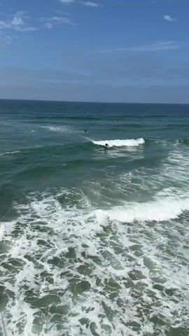 A surfer rides a wave in the ocean under a blue sky with scattered clouds. The camera is positioned above the water, looking down at the scene. The water is a deep green color, with white foam from the breaking waves. Several other surfers are visible in the distance.