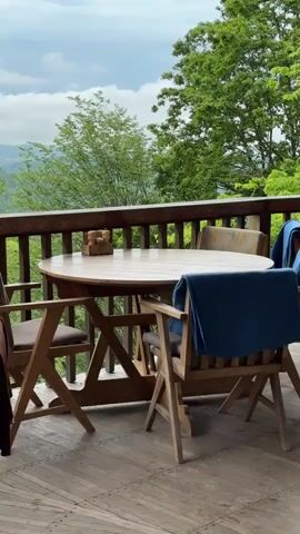 The video shows an outdoor wooden deck with a round wooden table and four chairs. Each chair has a blanket draped over its back. The deck has a wooden railing and overlooks a view of trees and distant mountains under an overcast sky. On the table is a wooden holder with salt and pepper shakers.  The flooring of the deck is wooden planks with alternating patterns.