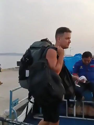 The video shows a man wearing a white t-shirt and black shorts with a large backpack and a black bag. He is walking on a boat and appears to be disembarking at a dock. There are other people on the boat and on the dock, as well as a truck and some blue tarps. The sky is overcast and the water is calm.