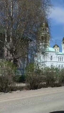 The video shows a white cathedral with multiple golden domes. The cathedral is located near a road, with a green fence separating it from the street. There are bushes and trees in front of the cathedral. The sky is blue with some clouds. The video is taken from a moving vehicle. There is music playing in the background. A red flag is also visible on the right side of the frame. There are cars parked near the cathedral.