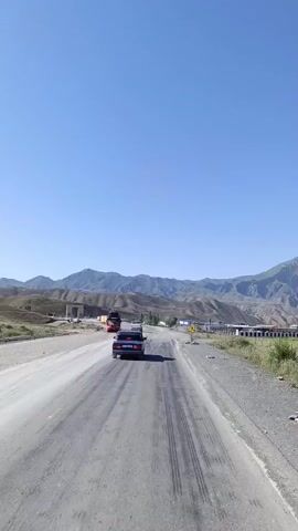The video shows a road with two lanes in a mountainous region under a clear blue sky. Cars are traveling on the road. A red truck is visible in the distance. There are mountains in the background. Traffic signs are posted along the side of the road, including a sign with a yellow triangle and a pedestrian symbol, and a circular sign with the number 50 inside. The road appears to be paved with asphalt but is also dusty and has tire marks. Green vegetation is growing on the side of the road, with a fence visible in some parts. The scene suggests a sunny day in a rural or semi-rural area.