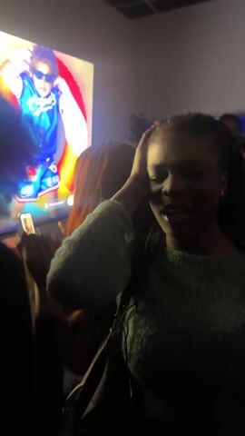 The video shows an indoor setting with multiple individuals. A large screen displays visual content, and the room appears to be somewhat dark with limited lighting. The individuals in the video are seen moving and interacting within the space. One person is holding a red cup. The ceiling has a grid pattern. The scene is somewhat chaotic.