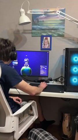 The video begins with a man sitting at a desk, playing a game on a computer. He is wearing a headset and using a mouse. The computer has a case with blue lights. There is a lamp, artwork, and a framed photo of a young girl above the desk. The camera quickly pans to another person, a young man with short brown hair and glasses, sitting on a bed. He has his hand behind his head. He looks toward the camera and smiles slightly.