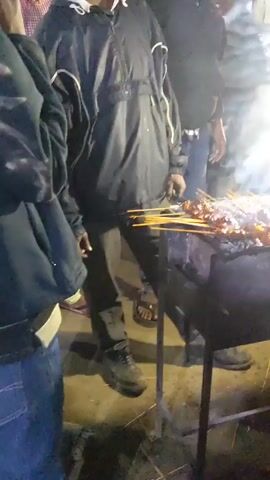 Here is a detailed description of the video:

The video shows a street food vendor grilling meat skewers over an open charcoal grill at night. Several people surround the grill, seemingly waiting to purchase the skewers. The grill is set up on a makeshift stand, and a bright light illuminates the food and the surrounding area. Various condiments and utensils are placed on a surface next to the grill. Smoke rises from the grill, adding to the atmosphere of a bustling street food scene. The people are dressed casually, suggesting an informal setting. The video captures the energy and activity of a nighttime street food market.