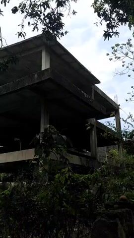 The video shows a dirt path leading through a wooded area. The path is lined on one side by a low, mossy brick wall and on the other side by trees. The trees overhang the path, creating a shaded canopy. The ground is covered in leaves. In the beginning of the video, the camera is pointed towards an unfinished concrete building that is overgrown with foliage. The camera then pans to the right to show the dirt path through the wooded area.