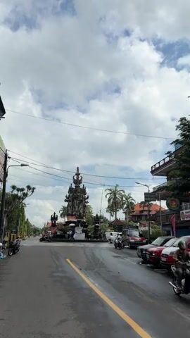 This vertical video showcases a bustling street scene in Klungkung, Indonesia, centered around a large, intricate monument. The monument, likely a significant cultural or religious landmark, is a towering stone structure adorned with multiple figures and decorative elements. It is draped with colorful fabric banners.

The scene is a street intersection or roundabout, with traffic actively flowing. Several vehicles, including cars and motorcycles, are visible navigating the area. A red scooter is prominently featured near the monument. On the right side of the road, buildings with traditional Indonesian architecture, featuring red roofs and balconies, line the street. One building has a sign that appears to have text in Indonesian.

The weather appears to be partly cloudy, with a mix of sun and scattered white clouds against a bright blue sky. The time of day seems to be daytime, likely late morning or afternoon, given the lighting conditions. The overall atmosphere suggests a lively, active urban environment. The wet pavement in the foreground may indicate recent rain. The presence of palm trees hints at the tropical climate of the location.