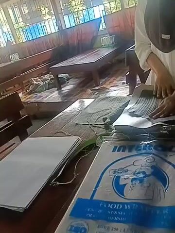 The video shows a close-up shot of a wooden table with various items on it. A person, wearing a hijab and a white long-sleeved shirt, is in the process of arranging some green leaves and white paper. In the foreground, a large plastic wrapper with the word "INTER" and "FOOD WRAPPER" is visible. The background suggests an indoor setting, possibly a classroom or a community hall, with wooden benches and tables, and windows with a view of greenery. The overall scene appears to be one of preparation or packaging.