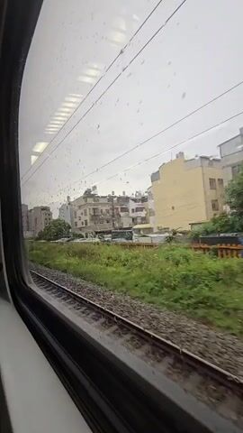 The video shows a view from a train window as it travels through a semi-urban area on a cloudy day. Raindrops are visible on the window. A railway track runs alongside the train, with lush green vegetation bordering it. In the distance, several multi-story buildings, some with balconies and air conditioning units, are clustered together. Cars are parked along a street in front of some of the buildings. Overhead power lines for the train are also visible. The overall impression is of a slightly overcast, rainy day in a built-up area.