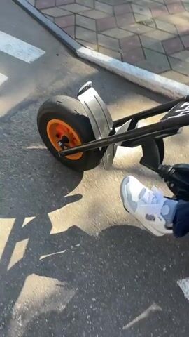 A man and a young girl are riding a go-kart on a paved path in a park. The man is in the front, driving, while the girl is seated behind him. They are both wearing casual clothing. The go-kart appears to be electric, with orange accents on the wheels and seats. The path is lined with trees and a decorative tiled area. The sun is shining, casting shadows on the ground, indicating it is likely a sunny day. The scene suggests a leisurely outing or a fun activity for the family. There is some text visible on the rear fender of the go-kart, but it is too blurry to be legible. The overall atmosphere seems pleasant and relaxed.
