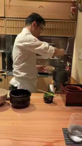 A man in a white chef's coat is preparing something behind a wooden counter. He is wearing glasses and has short black hair. He uses a ladle to pour liquid from a cream-colored bowl into a black bowl. On the counter, to his left, is a small, squat, black tea caddy, a light brown ceramic bowl, and a small green container holding chopsticks. In the background are wooden cabinets and stainless steel equipment. A glass of water is visible in the lower right corner.