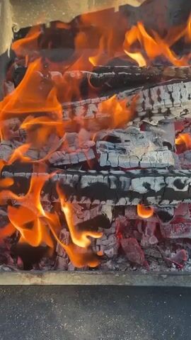 The video shows a close-up shot of burning logs in a fire pit. The logs are stacked horizontally and are engulfed in bright orange flames that lick at their charred surfaces. Embers glow red beneath the flames. The fire pit appears to be made of dark, rough material. The camera is positioned slightly above and to the side of the fire, focusing on the intensity of the flames and the texture of the burning wood.