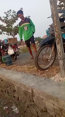 A young man in a green shirt and black shorts stands near a motorcycle on a dirt road. He smiles at the camera, then turns his head and puts his hand to his mouth, as if speaking. He then jumps onto a low stone wall and walks along it. In the background, another person on a motorcycle rides past. Further in the distance, there are trees and buildings. The scene appears to be outdoors during the daytime.