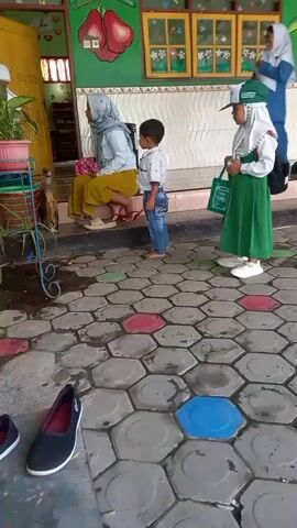 A group of children and adults are standing outside a building with colorful decorations. The building appears to be a school or daycare. There are plants in pots and shoes on the ground. The children are wearing uniforms, suggesting they are students. One child is being carried on another child's shoulders. An adult is sitting down and another is standing. The ground is paved with hexagonal tiles.