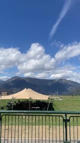 This video shows an outdoor scene with a large, tan, tension-fabric tent set up on a grassy field. Beneath the tent, there appear to be rows of chairs and some equipment, possibly for a performance or event. In the background, a majestic mountain range dominates the horizon under a bright blue sky dotted with fluffy white clouds. Several people are visible on the field in the distance, some appearing to be engaged in activities. A green metal railing is in the foreground, suggesting the viewer is looking from a slightly elevated or designated area. The overall impression is of a scenic outdoor gathering or event in a mountainous region.