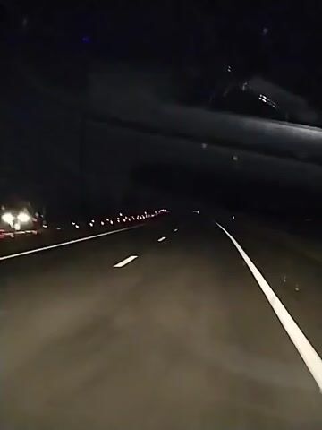 A first-person perspective video captures a car driving at night through a tunnel. The tunnel is dark with overhead lighting, and the road has white lane markings. The car's headlights illuminate the asphalt. The camera is positioned to capture the road ahead and the interior of the vehicle, including the dashboard. The motion of the car is evident, with the lane markings appearing to move and blur. The video is short and focuses on the experience of driving through the illuminated tunnel at night.