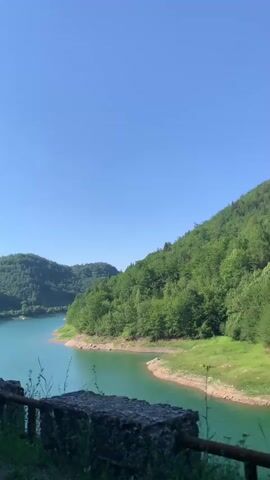 The video shows a view of a calm lake surrounded by lush green trees. The sky is clear and blue. The camera pans slightly to the right, revealing more of the tree-covered hills and the serpentine shape of the lake. There are some rocks and a rusty metal railing in the foreground. The water is a vibrant turquoise color.