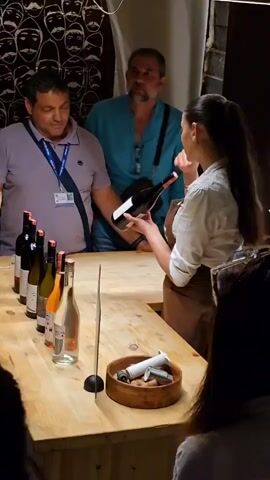 The video shows a woman, likely a sommelier, holding a bottle of red wine and addressing a small group of people. She is standing behind a wooden counter with several wine bottles lined up on it, including bottles with white, rosé, and red wine. She has long dark hair pulled back in a ponytail and is wearing a white shirt and brown apron. In front of her is a wooden bowl containing corks and a corkscrew. There are three men standing to her left and the back of a person's head to her right. One man is wearing a lavender shirt, another a teal button-down shirt, and the third is a young boy. The background is dark, with what appears to be a chalkboard with writing or a drawing on it. The woman seems to be explaining something about the wine, gesturing with the bottle in her hands.