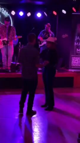 A bar scene is shown. On a stage, a four-man band is performing. A man playing an electric guitar is to the left, and a man playing the bass is to the right. In the foreground, a man and woman dance. The man wears dark pants and a gray shirt, while the woman wears dark pants, a dark top, and a beige hat. The stage is lit with red, white, and blue lights. A sign reading “JA MA” is on the right side of the stage. The man and woman hold hands as they spin around the dance floor.