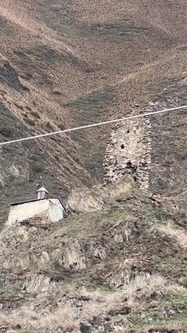The video shows a landscape scene featuring a small stone building with a dome-shaped roof next to the ruins of a stone tower. The buildings sit on a rocky, grassy hillside in front of a larger mountain covered in sparse vegetation. A power line stretches across the upper part of the image. The overall lighting suggests an overcast or cloudy day.
