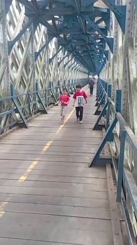 Two young boys and one adult male are walking and running on a wooden bridge with a blue metal superstructure. The bridge has a yellow line painted down the center of the walkway. The boy on the left is wearing a red t-shirt and light-colored shorts. The boy in the middle is wearing a white t-shirt with black lettering and dark pants. The adult male is further down the bridge, walking away from the camera. The bridge appears to be a pedestrian walkway and is enclosed by the metal structure. The environment outside the bridge is partially visible and appears to be green and leafy.