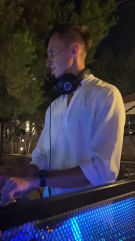 A medium shot shows a man wearing headphones and a white shirt standing behind a DJ setup. The man is looking down and his hands are on the DJ equipment. The DJ setup includes a laptop, a mixer with illuminated buttons and knobs, and two turntables. The background shows trees and some blurred lights, suggesting an outdoor night setting. The overall atmosphere is calm and focused.