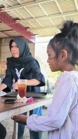 Here is a description of the video:

The video shows a covered outdoor dining area. A young girl in a striped shirt stands at a wooden table. A woman in a black hijab places plates of food on the table. Another woman in a hijab sits at the table, looking at her phone. The table also has glasses of iced tea, a cup of coffee, keys, and a phone charger. Other people can be seen in the background of the outdoor area.