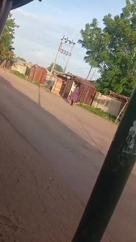 The video shows a street scene with a person walking away from the camera. The person is wearing a dark blue or purple robe and a head covering. They are walking on a dirt road next to a paved road. There are several small structures made of corrugated metal and wood along the side of the road. Trees and utility poles are also visible in the background. The lighting suggests it is daytime, possibly morning or late afternoon. The camera appears to be mounted on a moving vehicle, as indicated by the slight blur and angle.