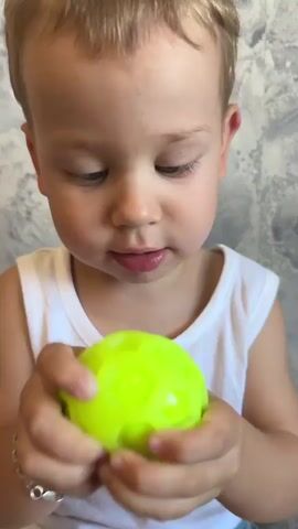 The video shows a young Caucasian child, appearing to be a toddler, sitting on a bed in an indoor setting. The child is wearing a white tank top and green patterned underwear. They have light brown hair and blue eyes. In the first frame, the child is close to the camera, with a bright yellow ball blurred in the foreground, suggesting it's being moved. The child's facial expression is slightly puzzled or curious, with pursed lips.

As the video progresses, the child is shown holding a textured yellow ball in their right hand, looking at it intently. The background is a textured wall, possibly wallpaper, with a grey and white pattern. The lighting suggests it is daytime, and the setting appears to be a home.

In the later frames, the child is now holding a small, neon green, multi-segmented toy in both hands and is examining it closely. The toy has a spherical shape with several protruding "bubbles" or segments. The child's focus is on the toy, and their expression is one of concentration. A silver bracelet is visible on the child's left wrist.

The overall activity is the child interacting with different toys, exploring them with curiosity and focus. The location is identified as Krasny Bor, Russia. There is no visible text in the video.