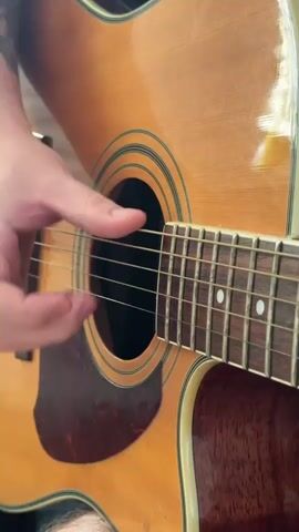 A close-up shot shows a person's hand playing an acoustic guitar. The hand is strumming the strings near the soundhole. The guitar is a light brown acoustic with a dark pickguard. The neck of the guitar, with its frets and fret markers, is visible. The background is blurred.