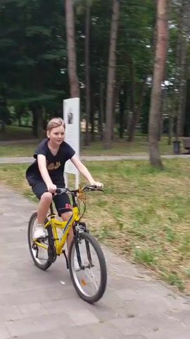 A person is riding a yellow bicycle down a paved path in a park. Trees line the path, and there are benches on either side. The person is wearing a black t-shirt and black shorts. They are looking forward and appear to be enjoying their ride.