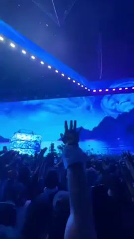 This video captures an energetic indoor concert or live event, showcasing a large crowd immersed in the experience. The setting is a spacious venue featuring a prominent stage with a massive LED screen and overhead lighting, all predominantly bathed in vibrant blue hues. The LED screen displays a dynamic, fantasy-like landscape, characterized by blue, cloudy skies, serene waters, and distant mountains, evoking a mystical or otherworldly atmosphere. Geometric, triangular light fixtures adorn the ceiling.

The crowd is highly engaged, with many people raising their hands in the air, some holding cell phones to record the performance. A brief close-up shot reveals two individuals smiling and appearing to sing along or cheer, further emphasizing the lively and enthusiastic mood of the event. The atmosphere is one of excitement and celebration. No distinct text is clearly visible on screen or in the crowd.
