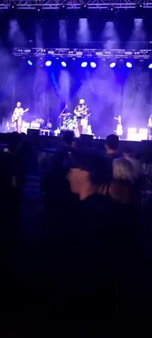 The video shows a live music concert with a band performing on stage. The stage is lit with bright white and blue lights, creating a dramatic effect. A crowd of people is gathered in front of the stage, watching the performance. Some members of the audience are visible in the foreground, including a person wearing a yellow shirt with "Josa Optics" printed on it. The band members are playing various instruments, including guitars and drums. The atmosphere appears lively and energetic, with the audience seemingly engaged with the music.