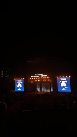 A crowd stands facing a concert stage. The stage has a large banner that says "Volanta World Concert Series" across the top. Two large screens flank either side of the center stage with blue displays and white text. There are bright orange lights over the stage. The video is shot from within the crowd.