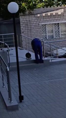 A man in blue overalls is bending over and working on a metal railing. He is standing on a step. There is a black cord with a coiled extension cord on the ground near him. To the left of the man is a black lamppost with a white globe light. The background consists of a brick building with windows and trees. The ground in front of the building is paved with gray brick.