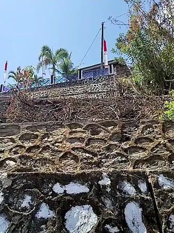 A low-angle shot pans across a textured stone retaining wall. Above the wall, a building with a blue tiled roof is visible, adorned with several Indonesian flags. Palm trees and other vegetation grow on the hill behind the building. The sky is clear and blue.