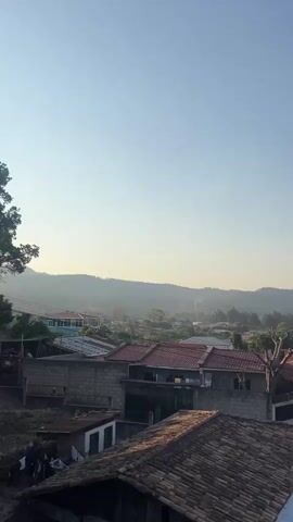 The video shows an elevated view of a neighborhood and surrounding mountains. The sky, initially clear, develops clouds as the video progresses. The neighborhood consists of closely packed houses with a variety of roof types. The mountains are visible in the distance, hazy and covered with trees. The camera pans slightly to the right to reveal more of the neighborhood. There are no people visible in the shot.