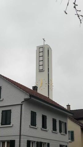 The video shows a church tower rising above the rooftops of a residential area on an overcast day. The tower is white and features a large clock face with gold-colored hands. A cross is perched at the very top of the tower. The buildings below have red-tiled roofs and green shutters on the windows. The sky is uniformly gray. The sound of church bells can be heard clearly. A small branch flutters slightly in the upper right corner of the frame.