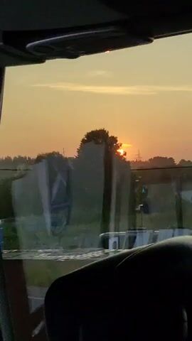The video captures a sunset through a windshield, presumably from inside a vehicle. The sun is a bright, orange orb low on the horizon, casting a warm glow across the sky. Silhouetted trees form a dark line against the colorful backdrop. In the foreground, a portion of the vehicle's interior is visible, including a dark seat and the edge of the windshield frame. Reflections on the glass show the outside environment, including what appear to be power lines and possibly some structures. The overall scene is serene and evokes the feeling of a peaceful end to the day.