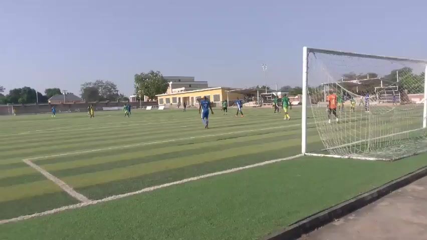 The video shows a soccer game being played on a green artificial turf field marked with horizontal stripes. The teams are wearing blue and green jerseys. There are players from both teams scattered across the field. The view is from behind the goal. Buildings are visible in the background, with trees and clear skies. The camera pans across the field showing the stadium seating.