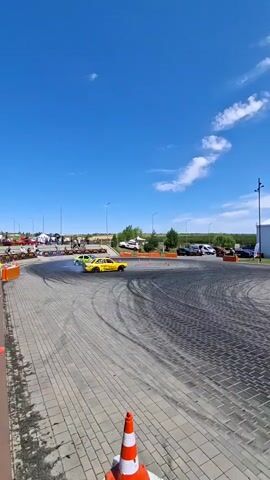 Two cars, one yellow and one lime green, are performing donuts on a paved lot. Both cars are leaving tire marks on the ground. In the background, there is a large white building with banners and a blue sky with white clouds. There are also orange traffic cones on the ground.