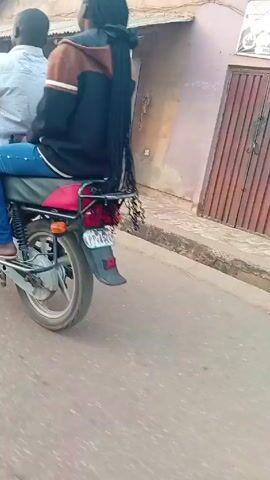 The video shows a red motorcycle with two passengers riding down a paved road in a residential area. The driver wears a light-colored shirt and tan pants. The passenger behind him wears jeans and a hooded jacket, with long braids hanging down their back. As the motorcycle travels, other motorcycles and pedestrians are visible on the sides of the road. Buildings and utility poles line the street, providing a backdrop for the scene. The overall setting suggests a small town or neighborhood.