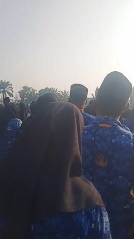 The video shows a group of people kneeling outdoors. The people are wearing blue uniforms with a repeating white pattern and some have black caps. Some individuals are wearing headscarves. The sun is bright and in the background, there are trees and a building. The video is shot from a low angle, looking up at the people. The overall impression is of a gathering or ceremony.