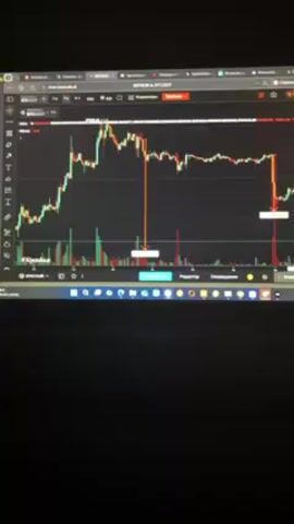 The video shows a laptop screen displaying a cryptocurrency chart, specifically of BTC/USDT, on a website titled "chart.krouskas.ai." The chart displays a series of red and green candlesticks, indicating price fluctuations over time. The screen is lit up, appearing in a dark room. The laptop is closed to a certain angle, with the lower part visible. The quality is not the best, causing a slight blur. Two orange vertical lines are placed on the chart, indicating specific time points or events on the chart.