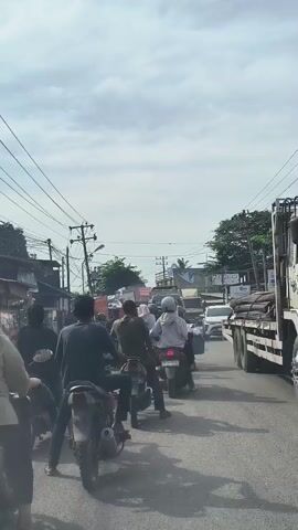 A vertical video shows a congested street with many motorcycles and cars. The sky is cloudy. The camera appears to be inside a vehicle, looking forward. Several people are riding on motorcycles, some wearing helmets and others not. One person in the foreground is wearing a white hoodie and a helmet. Traffic appears to be moving slowly. On the sides of the street, there are buildings and trees. There are also utility poles and overhead wires.