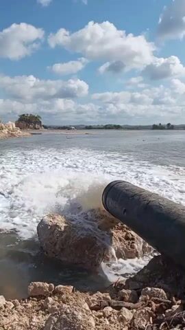A large pipe protrudes from the right side of the frame, discharging a powerful stream of foamy, murky water into a body of water. The water churns and splashes as it exits the pipe, creating a turbulent wake. The pipe appears to be made of dark, weathered material and is supported by rocks and earth on the shore. The shore itself is composed of loose soil, rocks, and debris. In the background, a vast expanse of water stretches to the horizon under a blue sky dotted with fluffy white clouds. A small island with vegetation can be seen in the distance. The overall scene depicts a forceful discharge of water into a natural environment.